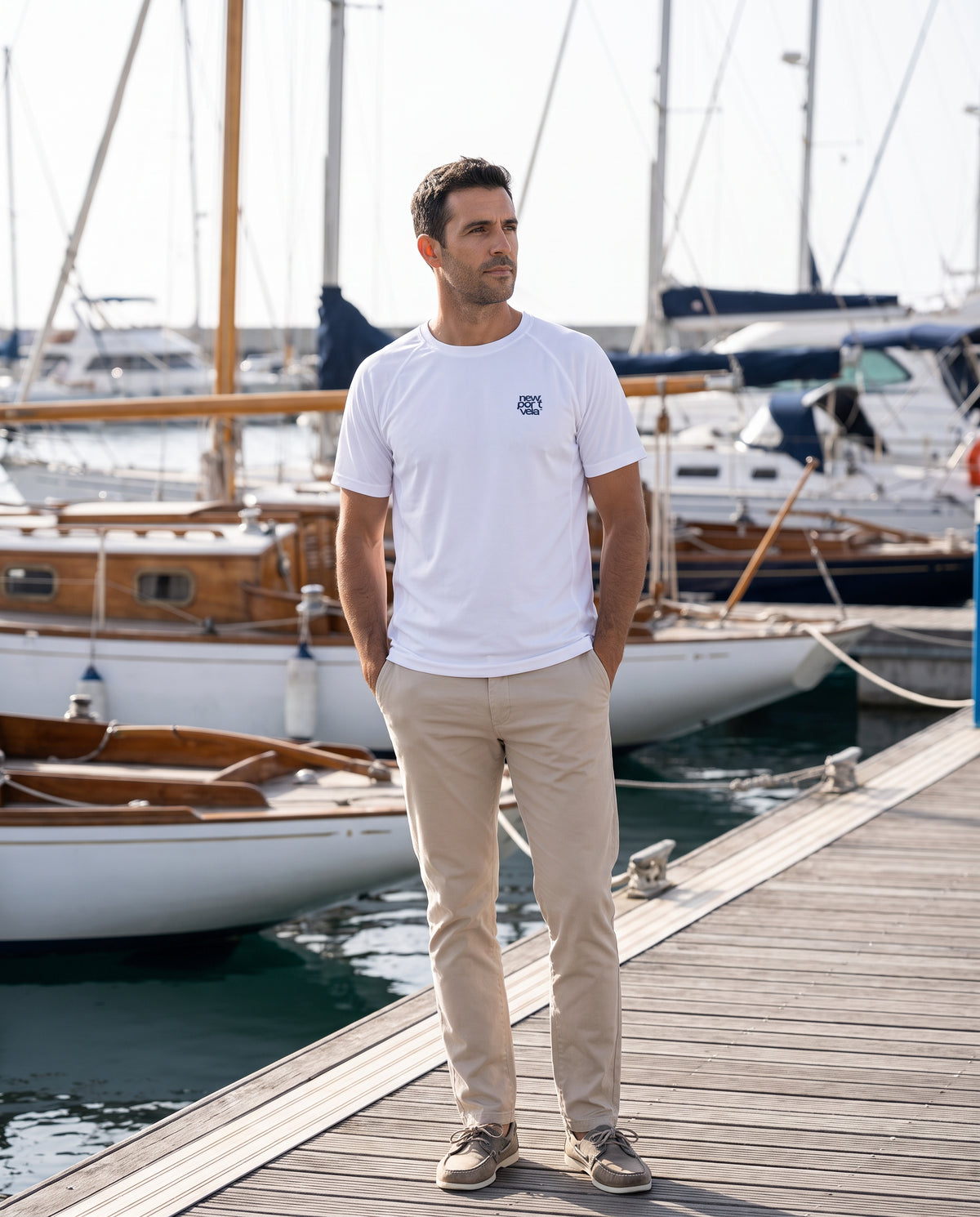 Man on a marina pontoon wearing the white NewPortVela Beaufort tech tee, with sailing boats moored behind