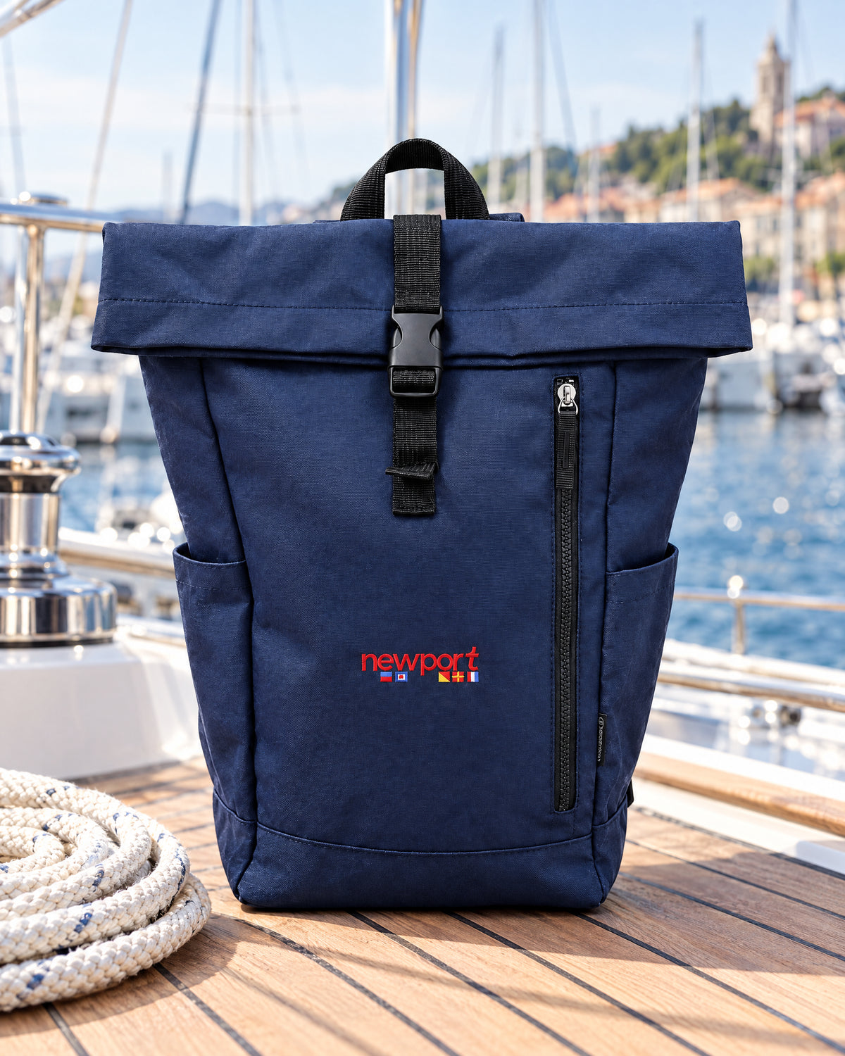 Navy NewPortVela Mironte roll-top backpack on the teak deck of a sailing yacht, with a coil of rope, the guardrail and the harbour behind